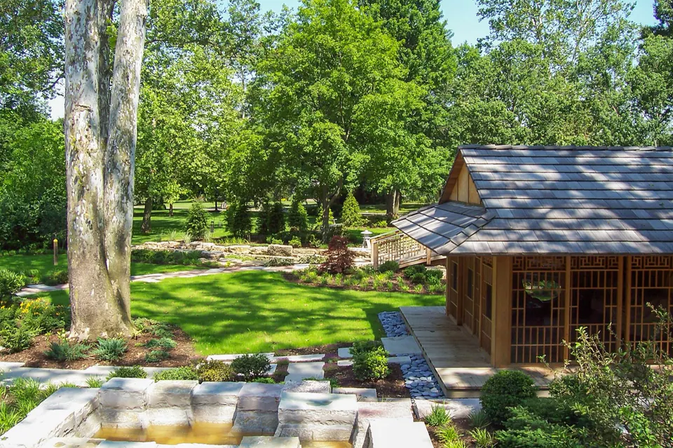 Wood garden pavilion beside large shade tree, lawn, bridge, stone path, and layered planting.